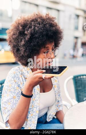 Junge afroamerikanische Frau, die ihr Telefon nahe ihren Mund bringt. Sie ist auf einer Barterrasse. Konzept der Sprachnachricht Stockfoto