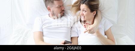 Wütender Mann, der das Smartphone seiner Frau in den Händen hält, während er auf dem Bett liegt Stockfoto