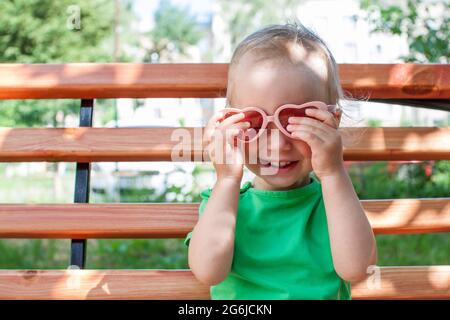 Das kleine Mädchen in einem grünen T-Shirt und einer rosa herzförmigen Sonnenbrille spaziert im Sommer im Park Stockfoto