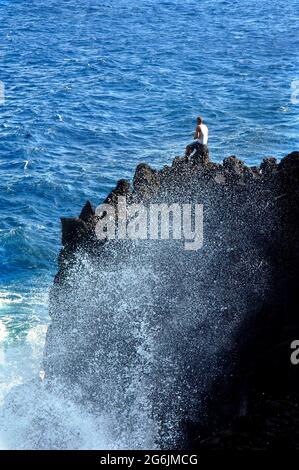 Der einheimische Fischer liegt am Rand der Klippe im McKinzie State Park auf der windwärts gelegenen Seite der Big Island. Eine große Welle stürzt ab und Spray erreicht h Stockfoto