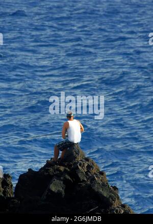 Ein einheimischer Fischer sitzt am Rande der Felsen im McKinzie State Park auf der Big Island von Hawaii. Er wird von der untergehenden Sonne hervorgehoben. Stockfoto