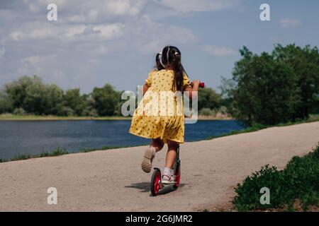 Lifestyle-Porträt Rückansicht eines Mädchens in einem gelben Kleid, das im Sommer auf einem Roller auf dem Flussufer reitet und ein Mädchen lernt, einen Roller zu fahren Stockfoto