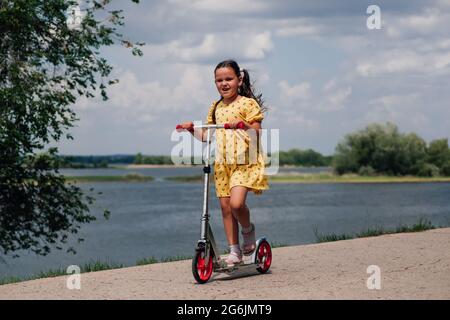 Glückliches, lächelndes Mädchen in einem gelben Kleid, das auf einem Roller reitet, hat eine gute Zeit und genießt einen Familienspaziergang entlang des Flusses an einem Sommertag Stockfoto