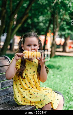 Ein süßes Mädchen in einem gelben Kleid sitzt auf einer Bank und isst im Sommer eine gelbe Maiskolben in einem Vergnügungspark Stockfoto