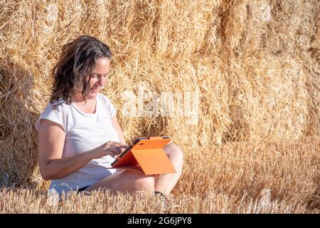 Die brünette Latina-Frau mittleren Alters sitzt auf dem Boden auf einem Feld und lehnt sich mit einer Tablette mit orangefarbenem Deckel auf einen Haufen Strohballen. Technologisch und Stockfoto