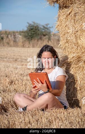 Die brünette Latina-Frau mittleren Alters sitzt auf dem Boden auf einem Feld und lehnt sich mit einer Tablette mit orangefarbenem Deckel auf einen Haufen Strohballen. Technologisch und Stockfoto