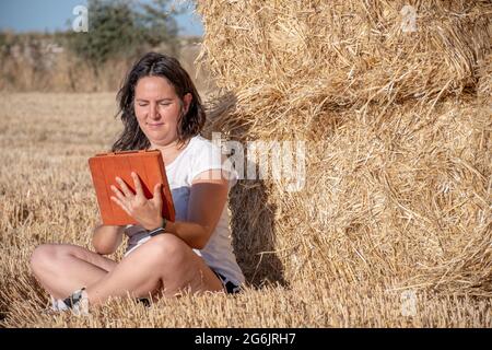 Die brünette Latina-Frau mittleren Alters sitzt auf dem Boden auf einem Feld und lehnt sich mit einer Tablette mit orangefarbenem Deckel auf einen Haufen Strohballen. Technologisch und Stockfoto