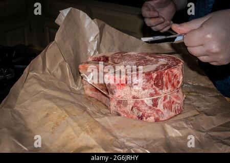 Rohes, zweibeinig geripptes Rindfleisch, das auf Fleischpapier gebraten wird, mit Pfeffer darauf, das von Hand mit Salz aus einem silbernen Ruder-Löffel gestickt wird Stockfoto