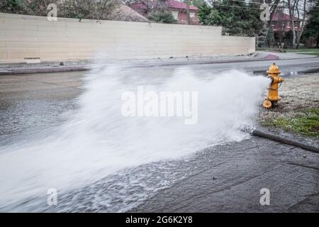 Gelber Hydrant an der Ecke der Kreuzung, der Wasser über die Straße mit großen Häusern und Bäumen im Hintergrund abgibt Stockfoto