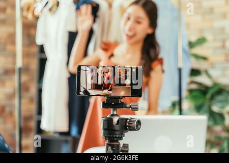 Junge und schöne asiatische Bloggerin zeigt Kleidung vor der Smartphone-Kamera, während sie Vlog-Video und Live-Streaming in ihrem Shop aufnimmt Stockfoto