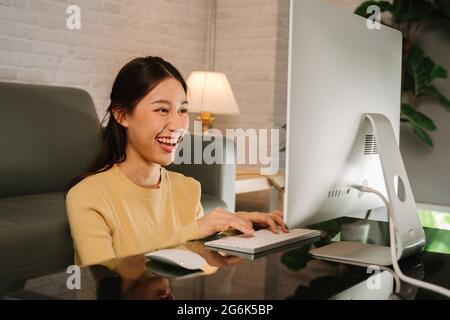 Erfolgreiche glückliche junge asiatische Geschäftsfrau, die von zu Hause aus arbeitet und mit einem Desktop-Computer auf der Tastatur tippt, während sie im Wohnzimmer sitzt Stockfoto