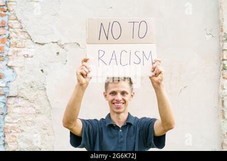 Der kaukasische Typ kam mit einem Plakat in den Händen heraus, um gegen Rassismus zu protestieren. Der Auftritt des jungen Mannes steht vor dem Hintergrund einer Zementwand mit Pappe in der Hand, auf der es Nein zum Rassismus heißt Stockfoto