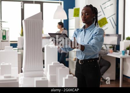 Architektin Frau afroamerikanischer Ethnie, die an Tablets arbeitet und ein professionelles Maquette-Baumodell ansieht. Architekturmitarbeiter betrachten Design für modernes Projekt in Entwicklung Stockfoto