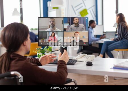 Behinderter gelähmter, ungültiger Mitarbeiter während eines virtuellen Meetings, der am Start-up-Büro über eine Webcam mit Partnern online diskutiert. Geschäftsfrau auf Videokonferenz. Stockfoto