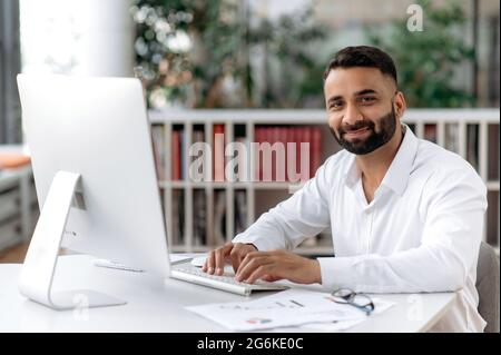 Porträt eines gutaussehenden, selbstbewussten, erfolgreichen bärtigen indischen Geschäftsmanns, Managers und Anwalts, der in einem weißen Hemd am Schreibtisch sitzt, direkt in die Kamera schaut und angenehm lächelt Stockfoto