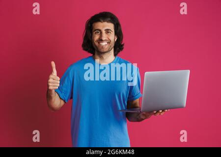 Unrasiert lächelnder Mann zeigt Daumen nach oben und hält Laptop isoliert auf rosa Hintergrund Stockfoto
