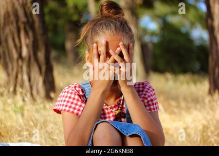 Kleines Mädchen versteckt Gesicht im Freien Porträt. Ein 5-10-jähriges Kind in einem Jeansshirt mit kariertem Hemd sitzt auf einem Feld, das das Gesicht mit den Händen bedeckt. Ein Kind Stockfoto