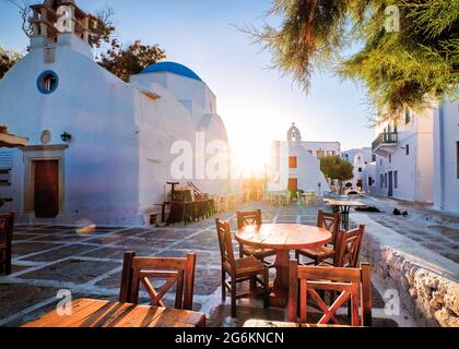 Sonnenaufgang in den Gassen von Mykonos, Griechenland. Weißgetünchte griechische Kirchen auf dem kleinen Stadtplatz, Straßencafé-Tische und -Stühle, Sonnen- und Linsenfackeln. Stockfoto