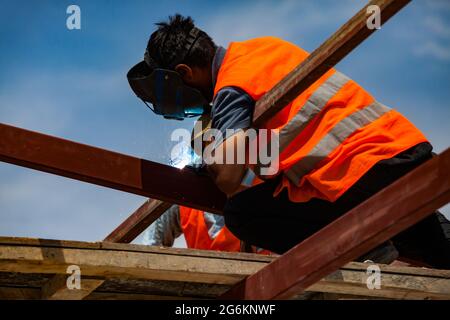 Nahaufnahme des Schweißers in Schweißmaske und orangefarbener Sicherheitsweste. Kein Gesicht. Flamme im Fokus. Anderer Teil des Fotos verschwommen. Blauer Himmel. Stockfoto