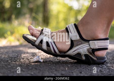Frau tritt in Sandalen auf einen verloren gegangenen kabellosen Kopfhörer, der auf einem asphaltierten Bürgersteig im Freien vor einem verschwommenen Hintergrund liegt. Stockfoto