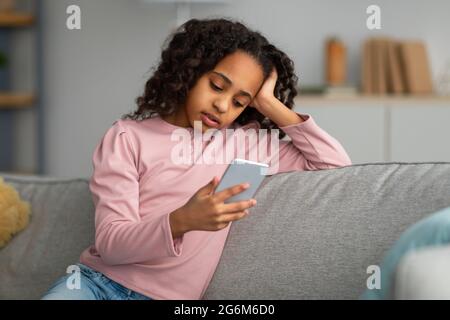 Gelangweilt African American Teen Girl Mit Smartphone Browsing Internet Sitzen Auf Sofa Zu Hause. Moderne Langweilige Kindheit Stockfoto