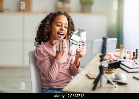 Schöne afroamerikanische Teenager-Bloggerin, die Lippenstift angesetzt und Spiegel angeschaut hat, während sie Vlog für ihren Kanal filmte Stockfoto