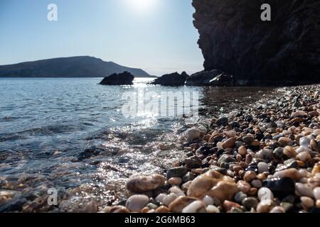 Die Sonne scheint auf dem wilden Kiesstrand aus nächster Nähe auf dem mittelmeer mit dunklen Felsen Silhouetten, klarem Wasser und Sonnenstrahl. Reisen Sie nach Griechenland in der Nähe von Athen. Sommer Stockfoto