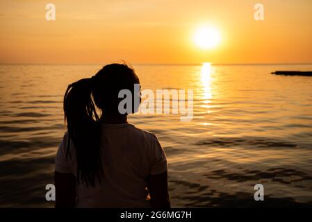 Frau, die den Sonnenuntergang am Meer betrachtet. Silhouette eines träumeren Mädchens, das hoffnungsvoll am Horizont schaut. Stockfoto