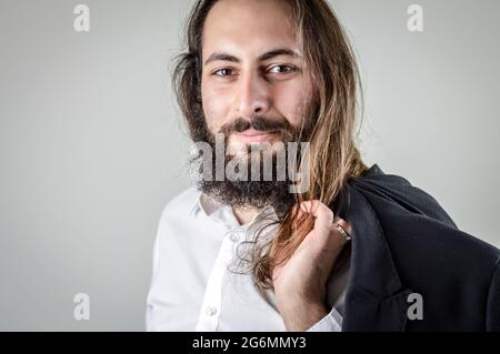 Porträt eines jungen Geschäftsmannes aus dem Nahen Osten mit Bart und langen Haaren, die seine Jacke über der Schulter halten Stockfoto