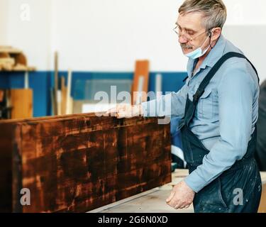 Ein grauhaariger Zimmermann mit Brille arbeitet mit Holz auf einer Werkbank in einer Schreinerei. Ein Rentner in einer medizinischen Maske bei der Arbeit. Beschäftigung und Arbeit ein älterer Mann in Arbeitskleidung Stockfoto