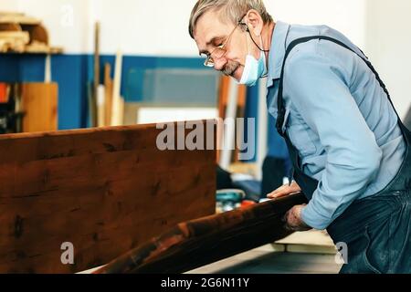 Ein grauhaariger Zimmermann mit Brille arbeitet mit Holz auf einer Werkbank in einer Schreinerei. Ein Rentner in einer medizinischen Maske bei der Arbeit. Beschäftigung und Arbeit ein älterer Mann in Arbeitskleidung Stockfoto