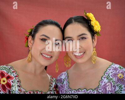 Schöne mexikanische Yucatecan Zwillingsschwestern mit Augen Make-up tragen traditionelles Yucatecan Outfit mit Blumen im Haar und lächeln für die Kamera. Stockfoto