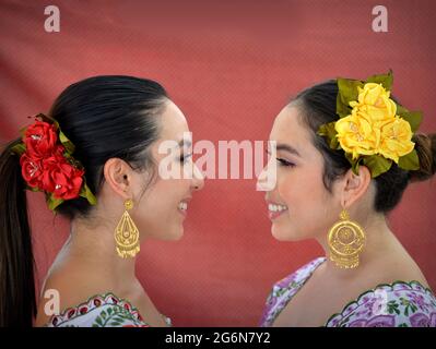 Schöne mexikanische Yucatecan Zwillingsschwestern mit Augen Make-up tragen traditionelles Yucatecan Outfit mit Blumen im Haar und sehen sich gegenseitig an. Stockfoto