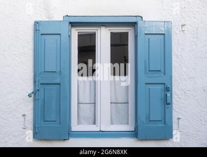 Traditionelles blaues Fenster, offene Holzläden und kleine weiße Vorhänge auf weißem Wandhintergrund. Die Fassade des weiß getünchten Gebäudes auf der griechischen Insel ist mit Details geschmückt. Cycl Stockfoto