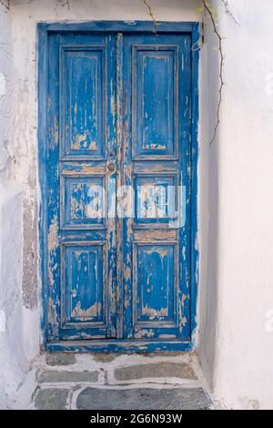 Blaue, alte, abgenutzte Tür auf weißem Wandhintergrund. Geschlossener, schäbiger Eingang aus Holz, Fassade des traditionellen kykladischen Gebäudes. Geschälte, verblasste Farbe. Griechische isla Stockfoto