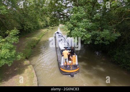 Crick, Northamptonshire, Großbritannien, 7. Juli 2021: Ein Paar mit einem kleinen Hund am Heck eines Schmalbootes segelt unter Bäumen entlang des Grand Union Canal. Stockfoto