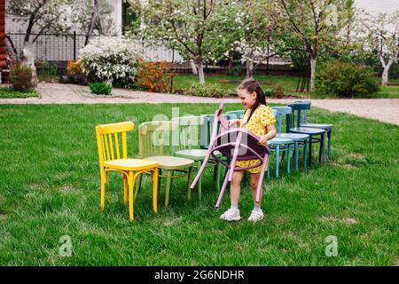 Ein Mädchen in einem gelben Kleid trägt einen Holzstuhl und arrangiert Stühle in einer Linie, um im Sommer im Garten eine Kindergeburtstagsfeier zu organisieren Stockfoto