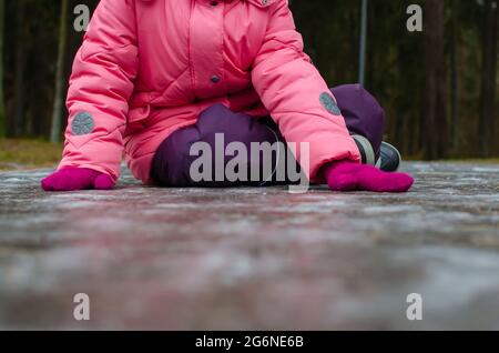 Ein Mädchen in einer rosa Jacke fiel in einen verschneiten Winterpark. Rutschte auf dem vereisten Weg. Gefahr von saisonalen Verletzungen. Stockfoto