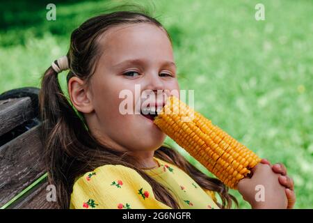 Ein glückliches fünfjähriges Mädchen knabbert an einem Sommertag auf einer Bank im Park Mais. Köstlicher gekochter Mais für Veganer Stockfoto