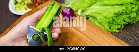 Der Prozess des Schälens einer Gurke aus der Nähe. Eine Frau in einer Schürze schält eine Gurke über einem hölzernen Schneidebrett. Kopfzeile der kulinarischen Seite Stockfoto