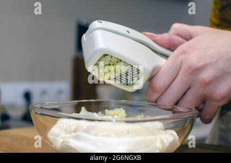 Knoblauchpresse aus Edelstahl und weißem Kunststoff. Knoblauchsoße in einer Glasuntertasse zubereiten. Stockfoto