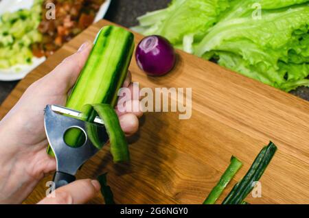 Der Prozess des Schälens einer Gurke aus der Nähe. Eine Frau in einer Schürze schält eine Gurke über einem hölzernen Schneidebrett. Stockfoto