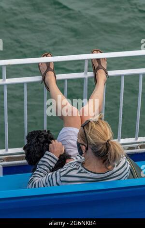 Frau, die auf einer Bank am Meer sitzt, mit einem kleinen Hund auf dem Schoß und ihren Füßen, die auf dem Geländer ruhen Stockfoto