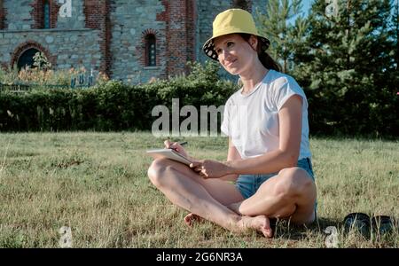 Mädchen sitzt im Park auf dem Gras und schreibt mit einem Stift in ein Notizbuch. Frau sitzt an einem sonnigen Tag auf dem Gras und schreibt in einem Notizbuch. Stockfoto