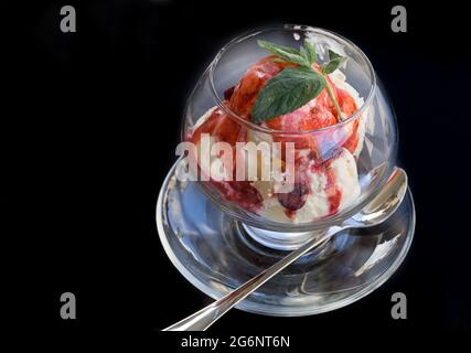 Vanilleeis mit Erdbeeren, Fruchtglasur und Minzblättern in einer Glasschüssel auf schwarzem Hintergrund. Speicherplatz kopieren. Stockfoto