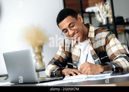 Smart positive Latino junger Mann mit Brille, sitzt am Tisch, mit einem Laptop, Notizen machen, studieren oder arbeiten online, hören ein Webinar, Wissen zu gewinnen, lächeln Stockfoto