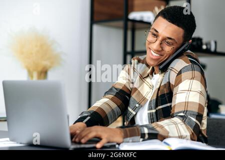 Beschäftigt positive Latino modernen erfolgreichen Kerl, ceo oder Designer, sitzen am Schreibtisch im Büro, mit einem netten Handy-Gespräch während der Arbeit mit Laptop, lächeln freundlich. Mobiltelefonkommunikation Stockfoto
