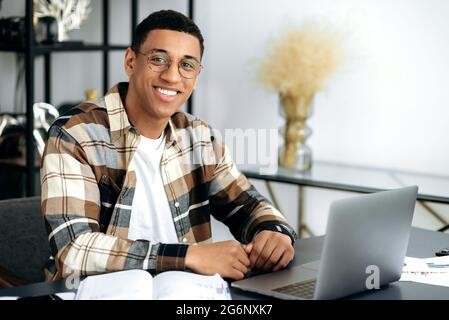 Porträt eines gutaussehenden, charismatischen, positiven hispanischen Mannes mit Brille, stilvoll gekleidet, Designer oder Freiberufler, der an seinem Schreibtisch im Büro sitzt, die Kamera anschaut und freundlich lächelt Stockfoto