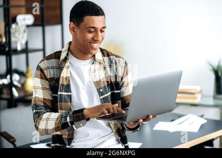 Positiv selbstbewusster, moderner, gemischter Latino-Mann mit Brille, kreativer Designer oder Freiberufler, stilvoll gekleidet, neben einem Schreibtisch mit offenem Laptop stehen, auf den Bildschirm schauen, lächeln, im Internet surfen Stockfoto