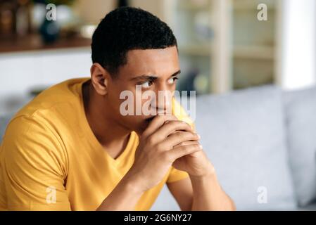 Frustrierter, unglücklicher, tausendjähriger Latino-Mann in einem gelben T-Shirt, der auf dem Sofa im Wohnzimmer sitzt, hat persönliche Probleme, Misserfolge bei der Arbeit, Depressionen, schlechte Laune, schlechte Nachrichten, Trauriger junger Mann ist einsam Stockfoto